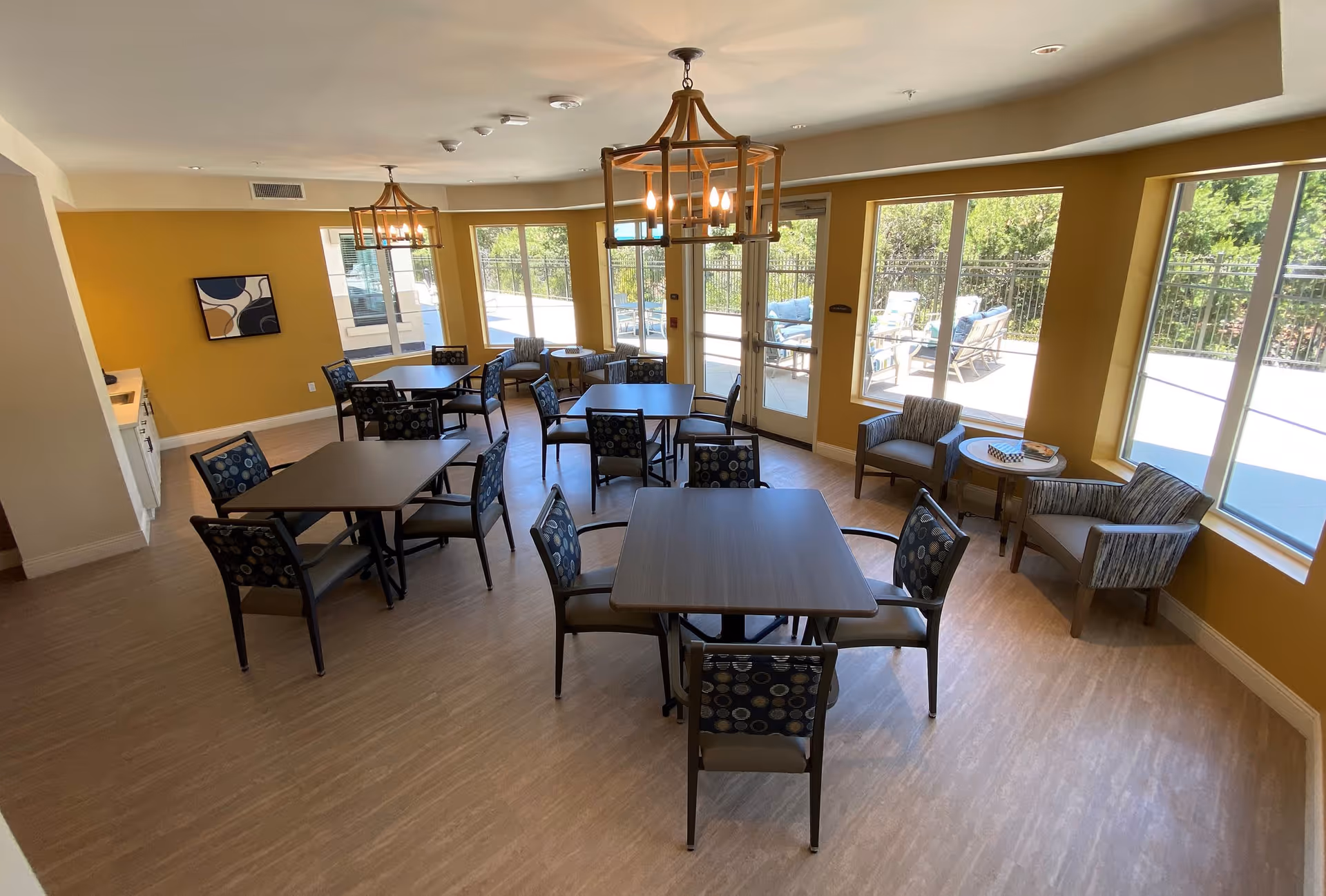 A bright dining room with several square tables and chairs arranged neatly. The walls are painted yellow, and large windows allow natural light to fill the space. There are two modern chandeliers hanging from the ceiling. Outside the windows, there is a patio area with outdoor seating visible.