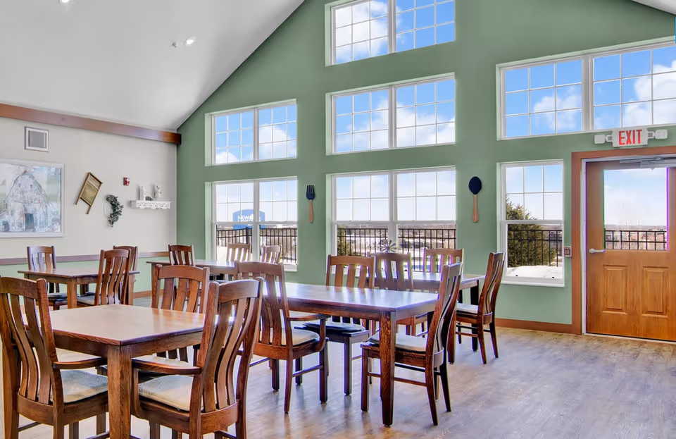Dining room with wooden tables and chairs beneath large multi-pane windows and a wooden exit door.