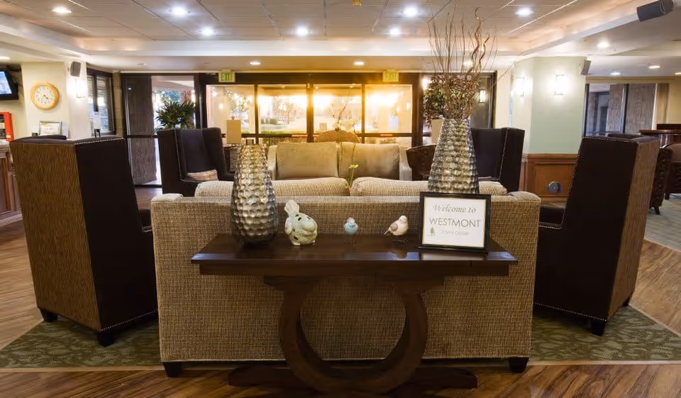 A cozy and well-lit common area in a senior living facility with beige sofas, dark brown armchairs, a wooden table with decorative vases and small figurines, and a framed sign that says 'Welcome to Westmont'. Large windows in the background let in warm light.