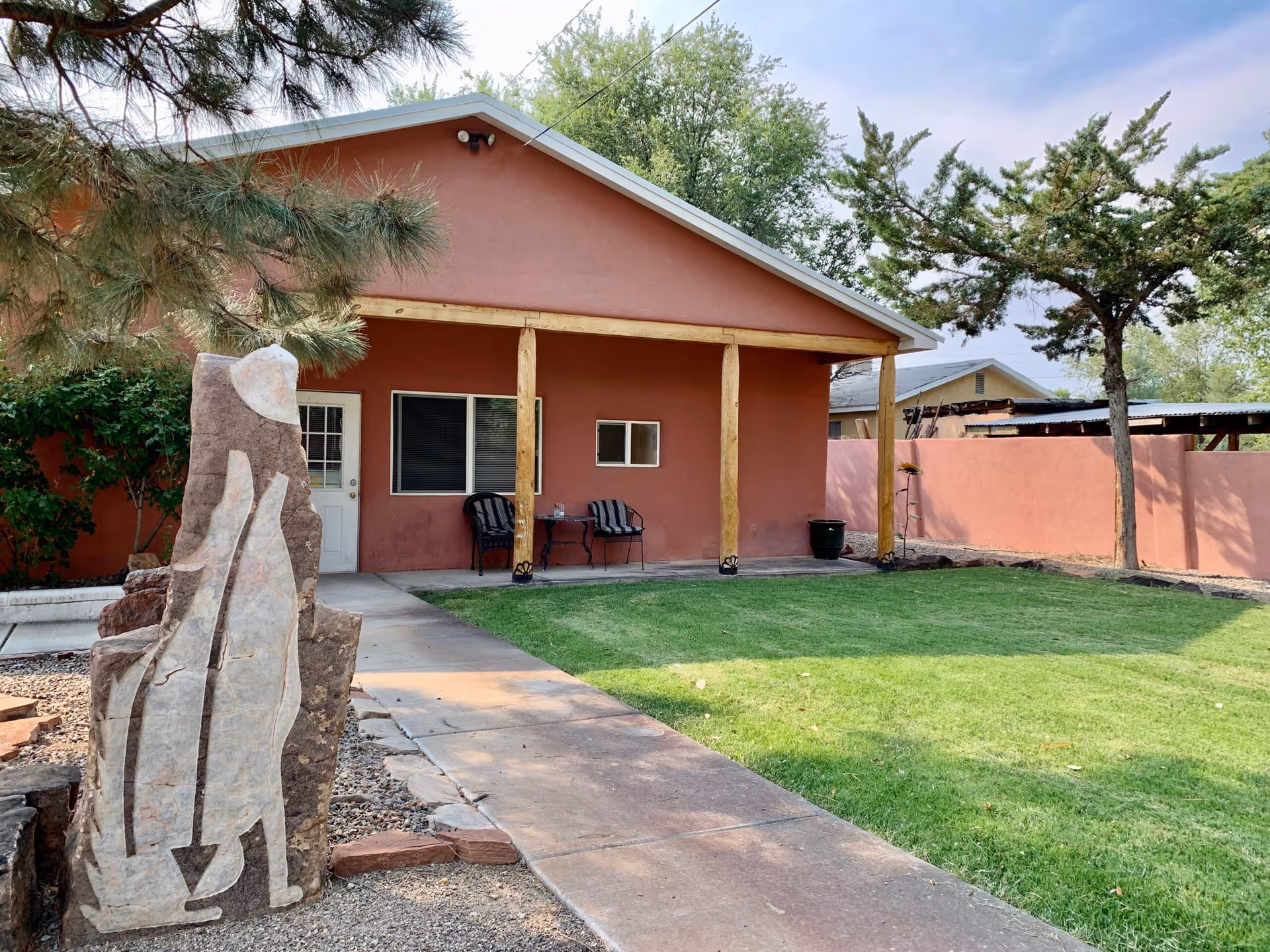 Outdoor view of Casa de Rosa Assisted Living showing a single-story building with a covered porch supported by wooden posts. There are two chairs and a small table on the porch. In the foreground, there is a stone sculpture and a concrete walkway leading to the porch. The area has green grass, trees, and a reddish-brown wall surrounding the yard.