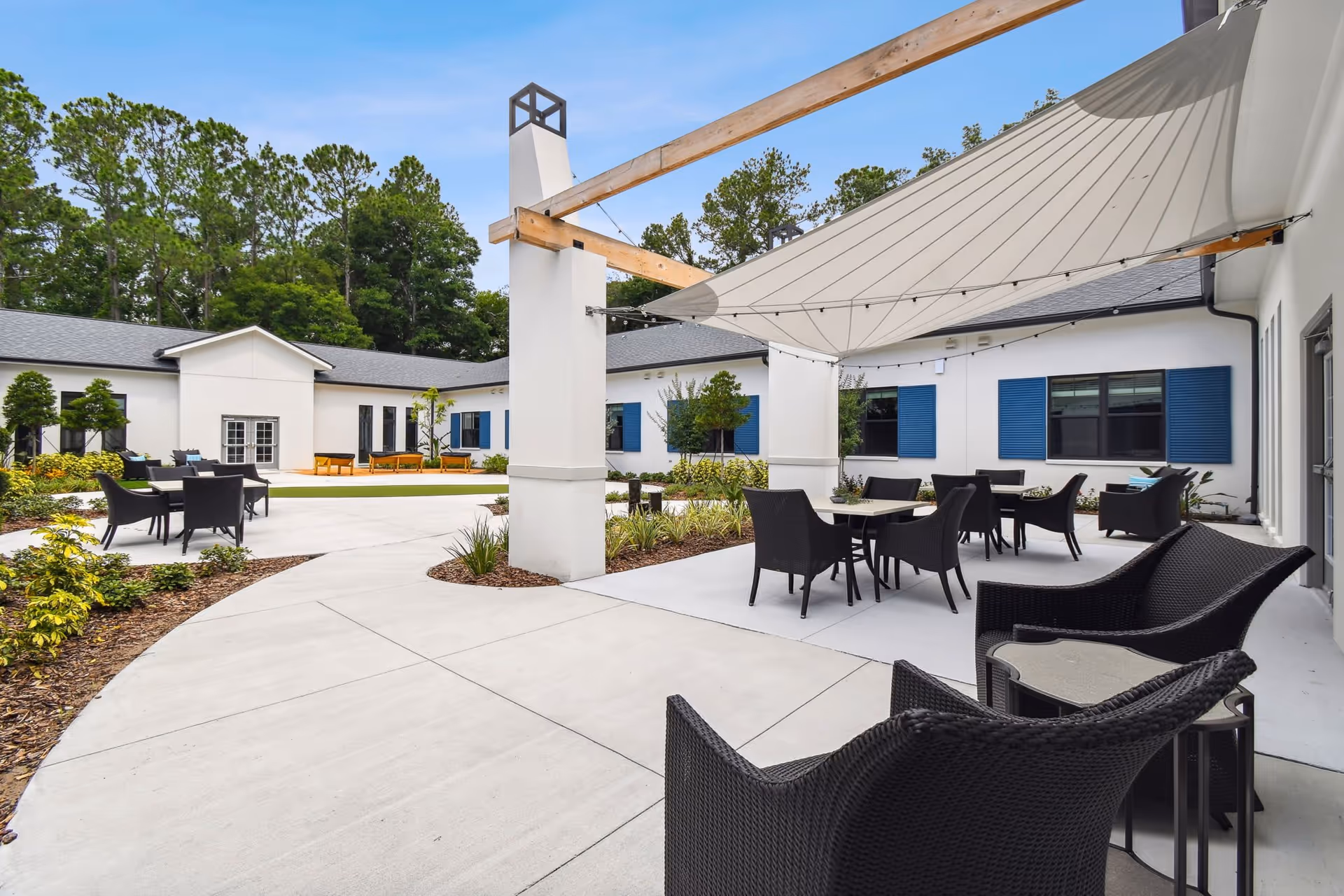 Outdoor courtyard area at Aden Senior Living featuring multiple black wicker chairs and tables under a large white shade sail, surrounded by a white building with blue window shutters and greenery.