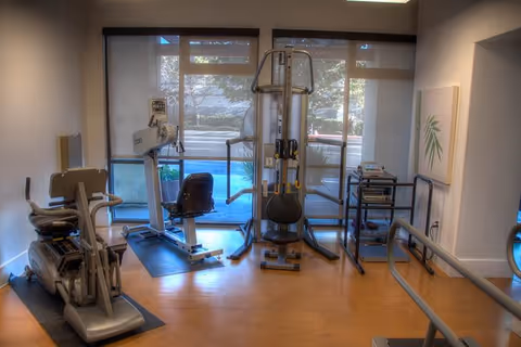 A small fitness room with exercise equipment including a recumbent bike, a multi-function weight machine, and a treadmill. The room has large windows with a view of the outdoors, light-colored walls, and wooden flooring.