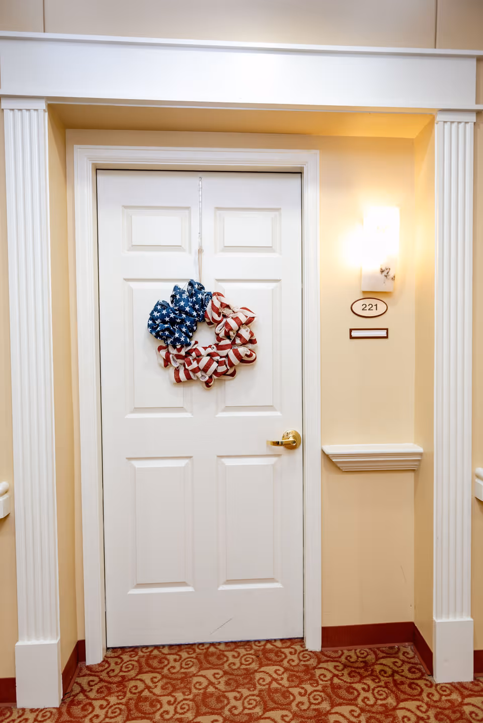 White apartment door in a hallway decorated with an American-flag wreath and labeled room number 221.