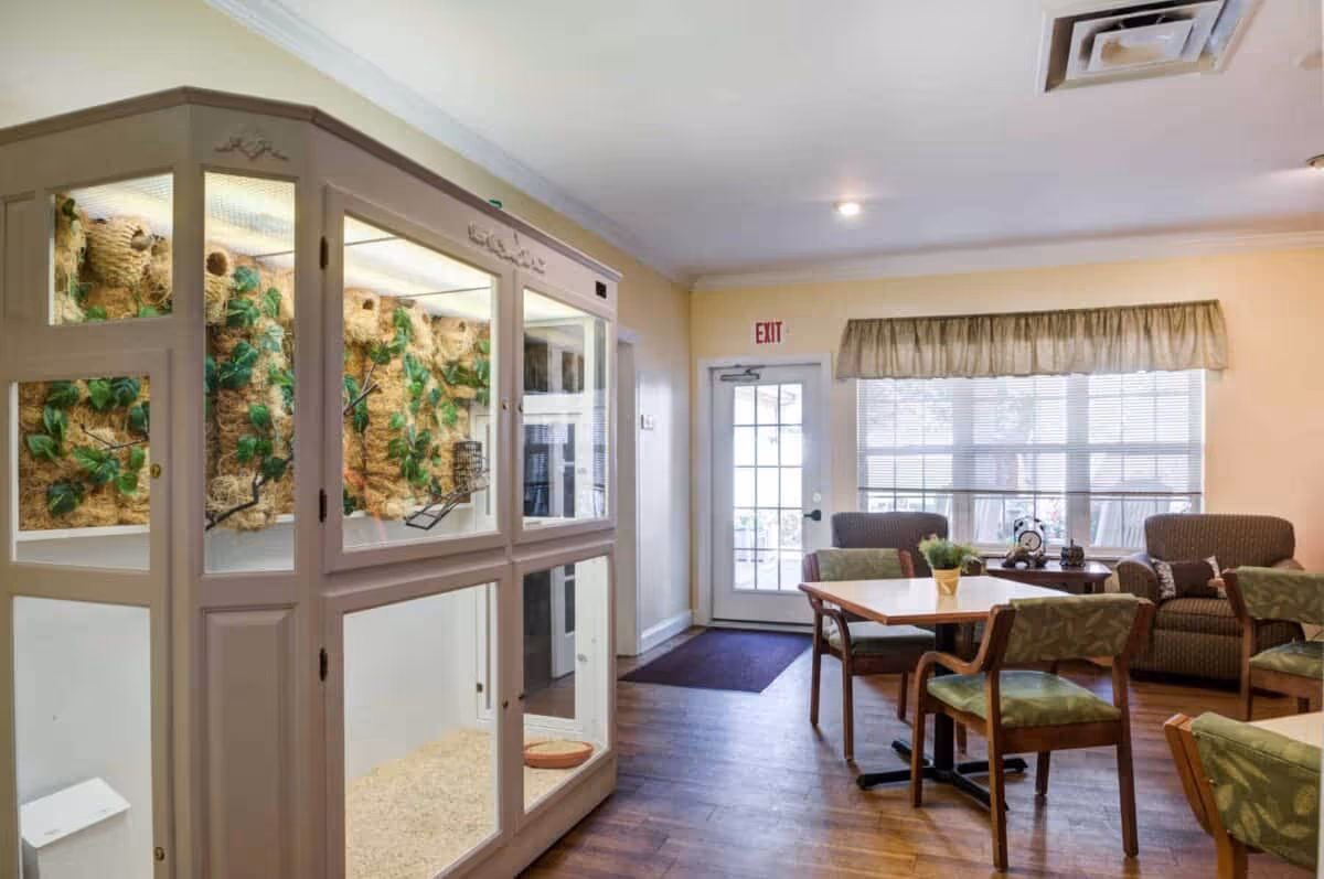 Interior view of a senior living facility common area with wooden flooring, a large glass-enclosed bird habitat on the left, several chairs and tables arranged for seating, a couch, and a door with an exit sign above it leading outside. The room is well-lit with natural light coming through a large window with blinds and a valance.