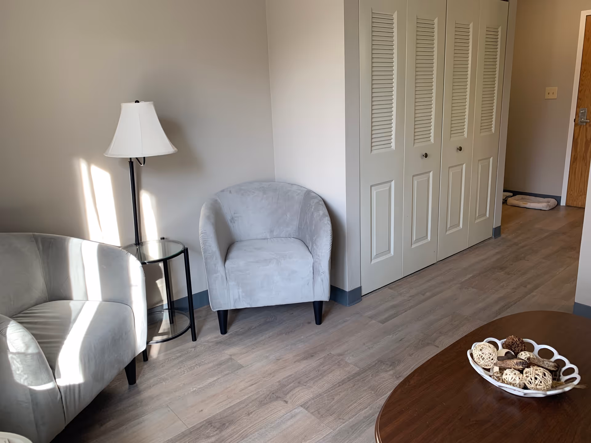 A cozy corner of a room with two light gray upholstered armchairs, a small round glass side table with a black metal frame, and a floor lamp with a white lampshade. There is a wooden coffee table with a decorative bowl filled with natural elements like pine cones and woven balls. The room has light wood flooring, beige walls, and white closet doors with louvered panels. A wooden door is visible in the background.