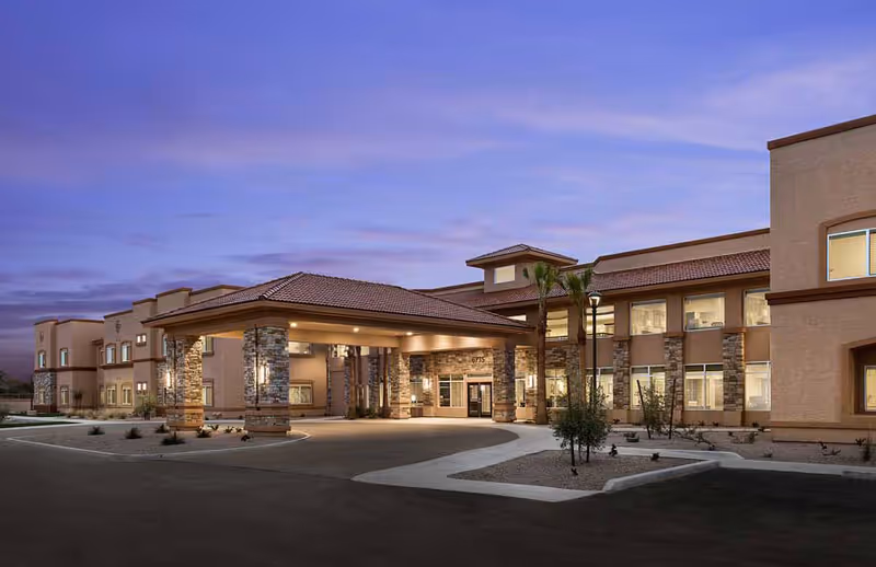 Exterior view of MorningStar Senior Living at Golden Ridge building at dusk, showing a two-story structure with stone and stucco facade, a covered entrance with pillars, palm trees, and a curved driveway.