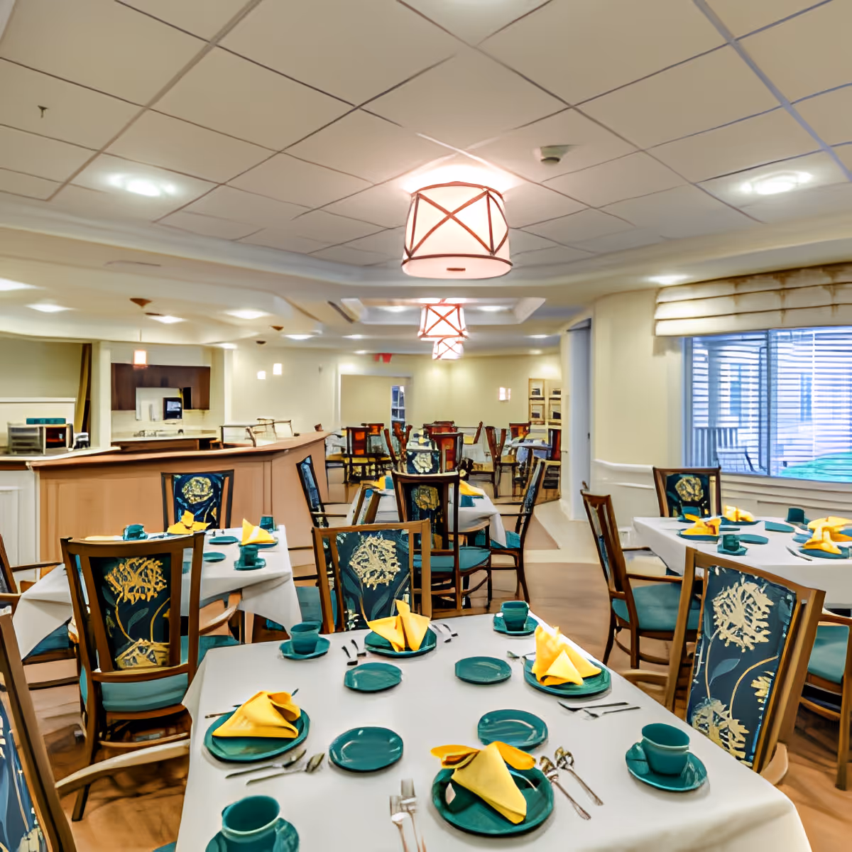 A dining room in a senior living facility with multiple tables set for a meal. Each table has white tablecloths, green plates, cups, and yellow folded napkins. The chairs have floral patterned upholstery. The room has soft lighting with ceiling fixtures and a large window with blinds on the right side.