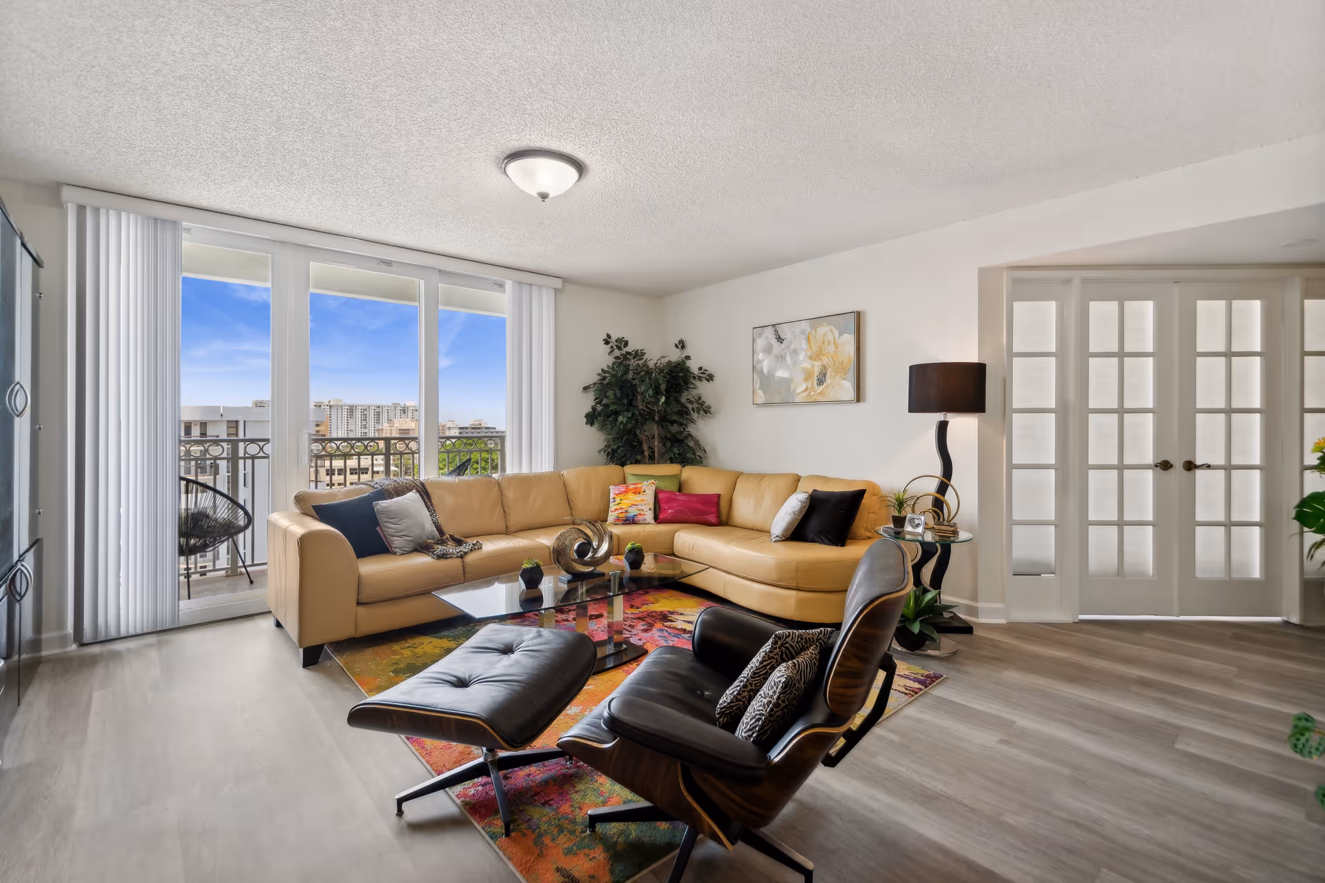 Bright living room with a tan sectional sofa, black lounge chair and ottoman, glass coffee table on a colorful rug, and sliding glass doors opening to a balcony with a city view.