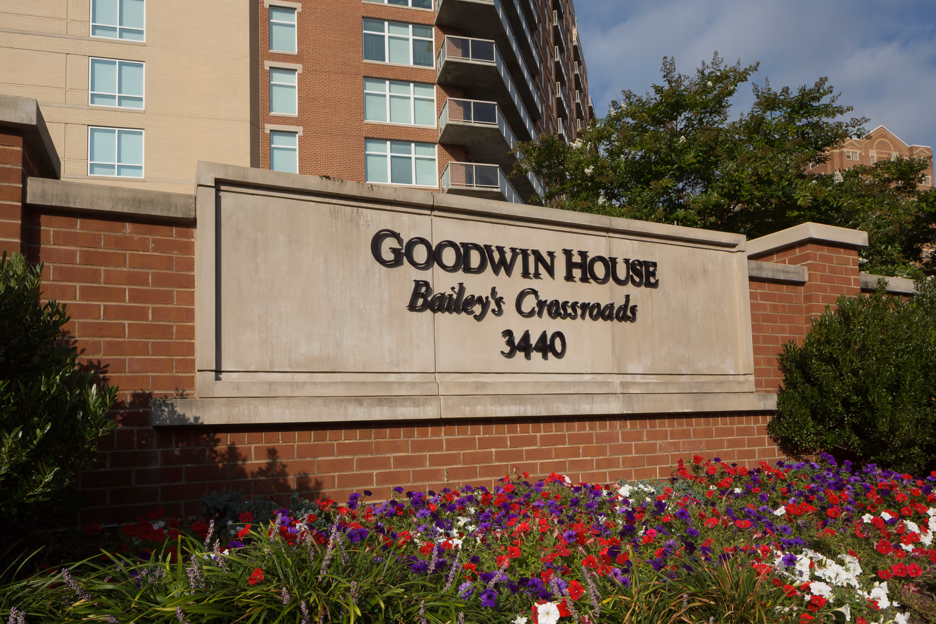 Outdoor view of a brick and stone sign for Goodwin House Bailey's Crossroads with the number 3440. The sign is surrounded by colorful flowers and greenery, with a multi-story building and trees in the background under a partly cloudy sky.