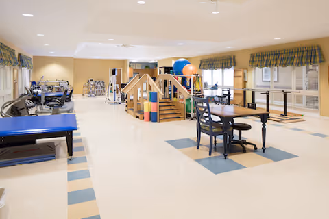A spacious therapy or rehabilitation room with exercise equipment, therapy balls, parallel bars, and a wooden ramp structure. There is a table with chairs in the center, and the room has large windows with plaid valances allowing natural light to enter.
