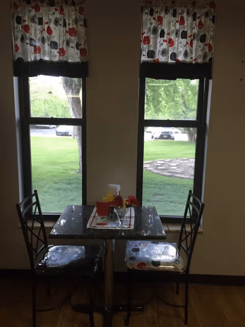 A small dining area with a square table and two chairs positioned in front of two tall windows. The windows have floral-patterned valances with red, black, and white colors. Outside the windows, green grass, trees, and parked cars are visible. The table has a decorative runner, a small vase with red flowers, and a container with napkins.