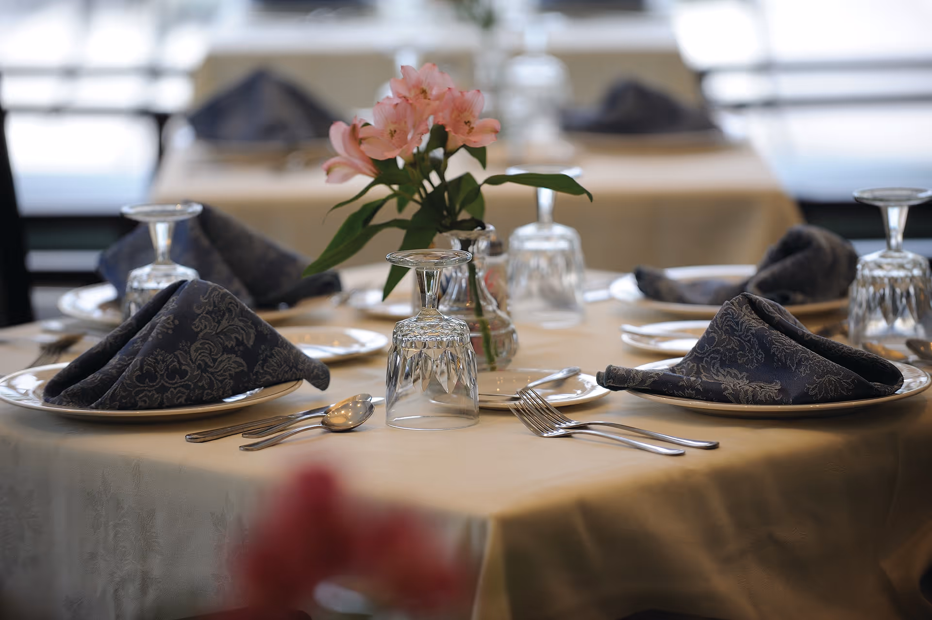 A close-up view of a dining table set with folded dark patterned napkins on white plates, upside-down clear drinking glasses, silverware, and a small vase with pink flowers in the center. The background shows more similarly set tables.