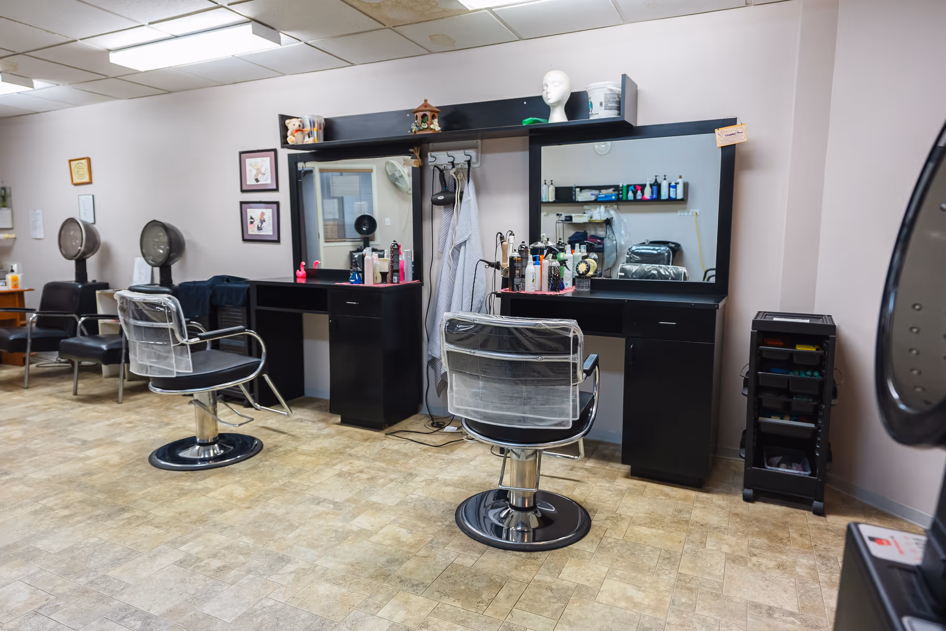 Interior view of a hair salon area in a supportive living facility featuring two black salon chairs with protective covers in front of large mirrors and black countertops. Various hair care products and tools are arranged on the counters. In the background, there are two hair dryers and framed pictures on the wall. The floor is covered with beige tiles.