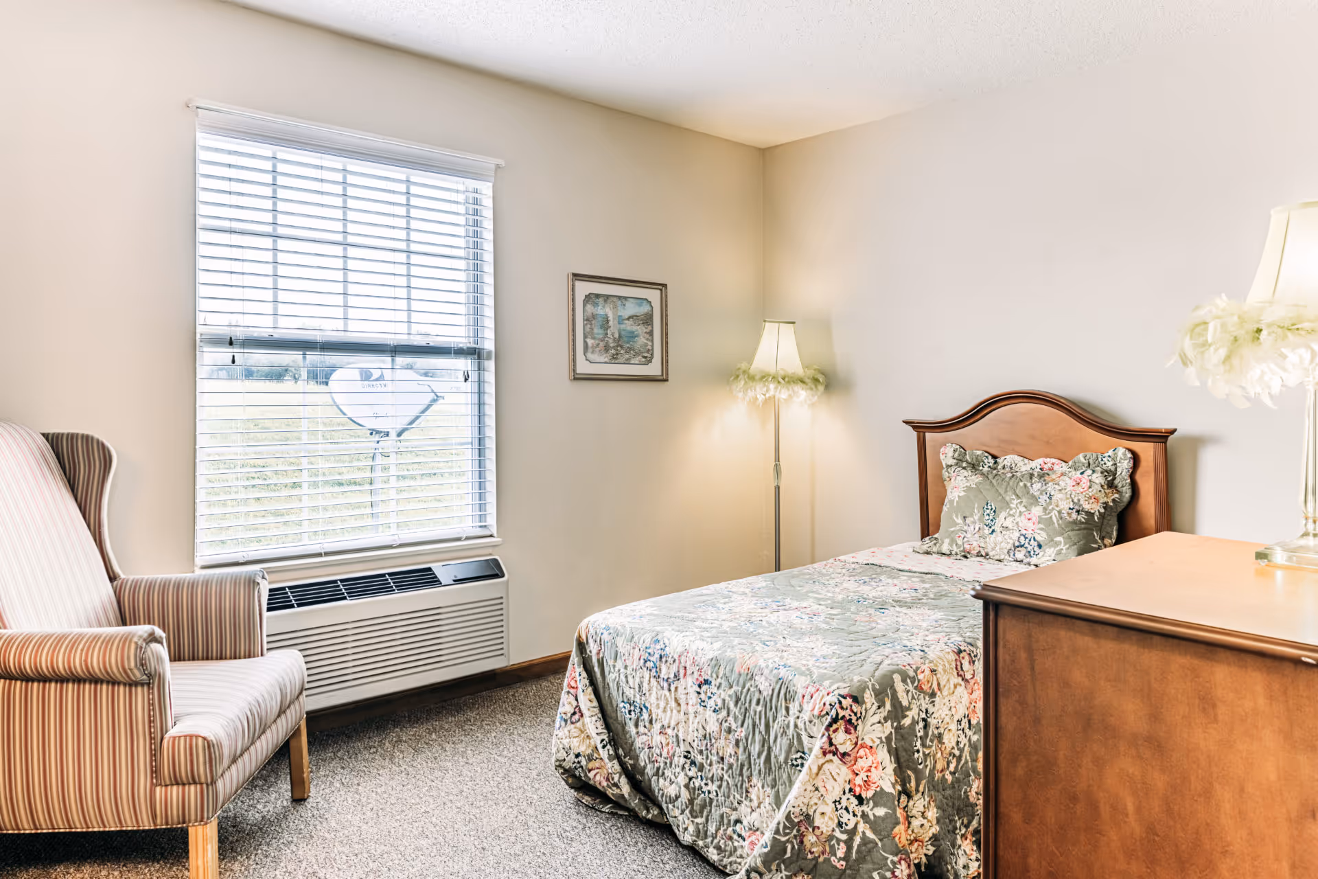 A cozy bedroom with a single bed covered in a floral quilt and matching pillow. Next to the bed is a wooden dresser with a lamp on top. A striped armchair is positioned near a window with white blinds, and a floor lamp with a feathered shade stands in the corner. A framed picture hangs on the wall.
