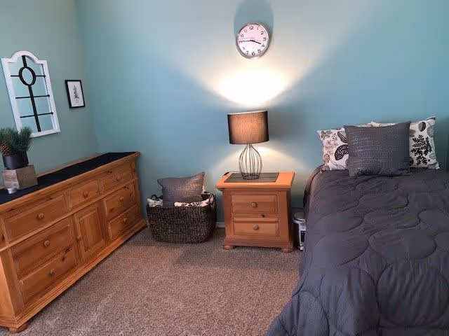 Neat bedroom with teal walls, a wooden dresser and nightstand, a lamp under a wall clock, a woven basket, and a bed with gray bedding.