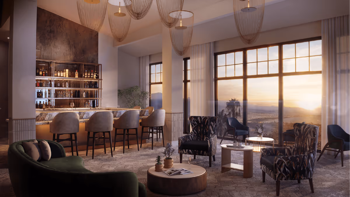 A stylish lounge area with a bar counter featuring several bottles of liquor on shelves. The bar has five cushioned bar stools. The seating area includes a green curved sofa, two patterned armchairs, and several small round tables. Large windows offer a scenic view of a sunset over mountains and palm trees. The room is softly lit with hanging pendant lights and has a cozy, elegant atmosphere.