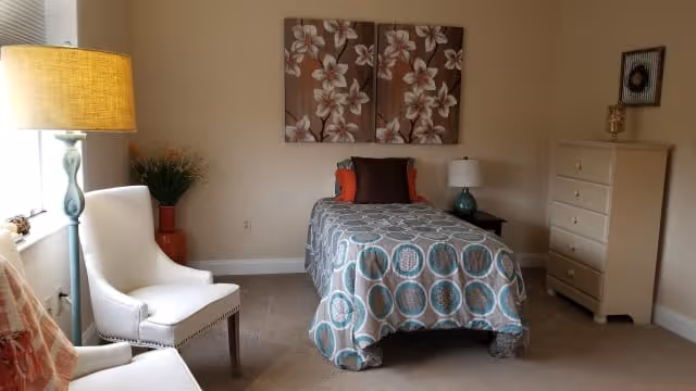 A cozy bedroom featuring a single bed with a patterned bedspread and multiple pillows. There are two white upholstered chairs, a tall lamp with a yellow shade, a potted plant in a red vase, a white chest of drawers, and floral artwork hanging on the beige wall.