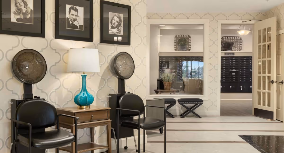 Interior view of a senior living facility hair salon area with two black salon chairs and hair dryers, a wooden side table with a turquoise lamp, and three black and white framed portraits on the wall. In the background, there is a window showing a room with mailboxes and two black stools.
