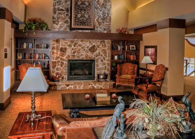 Cozy living room with a large stone fireplace in the center, flanked by built-in wooden bookshelves filled with books and decorative items. The room features comfortable upholstered armchairs, a leather sofa, a wooden coffee table with decorative items, and a table lamp on a side table. There are also two peacock sculptures and a plant arrangement on the coffee table.