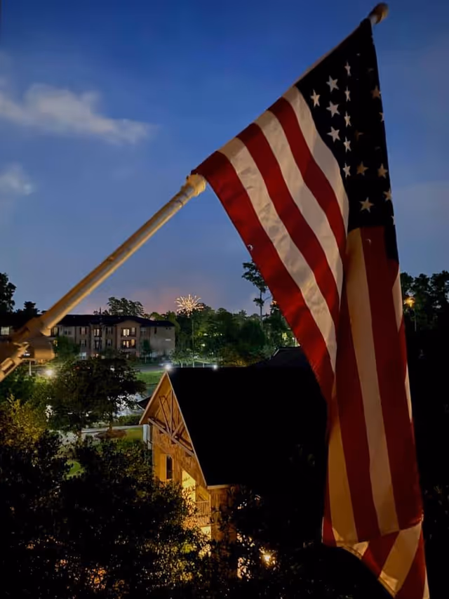 An American flag is prominently displayed on a flagpole in the foreground during dusk. In the background, there are residential buildings, trees, and a small building with warm lights. Fireworks are visible in the distance against a darkening sky.