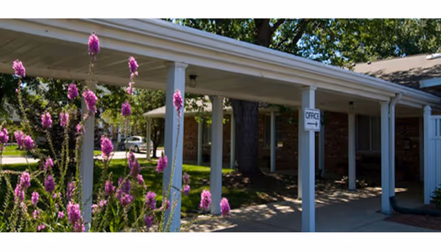 Exterior view of a single-story brick building with a covered walkway supported by white columns. Purple flowers are in the foreground, and a sign indicating the office direction is attached to one of the columns. Trees and greenery surround the area.