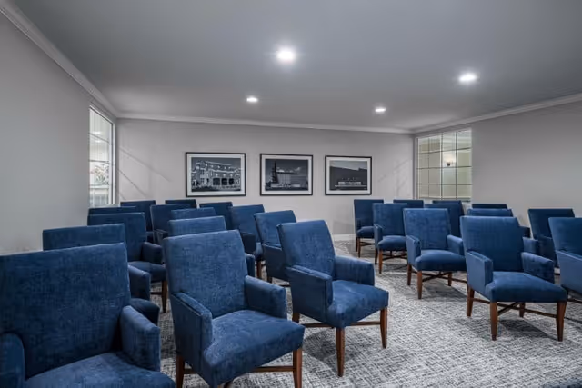A small meeting room with multiple blue upholstered chairs arranged in rows facing framed artwork on a gray wall.