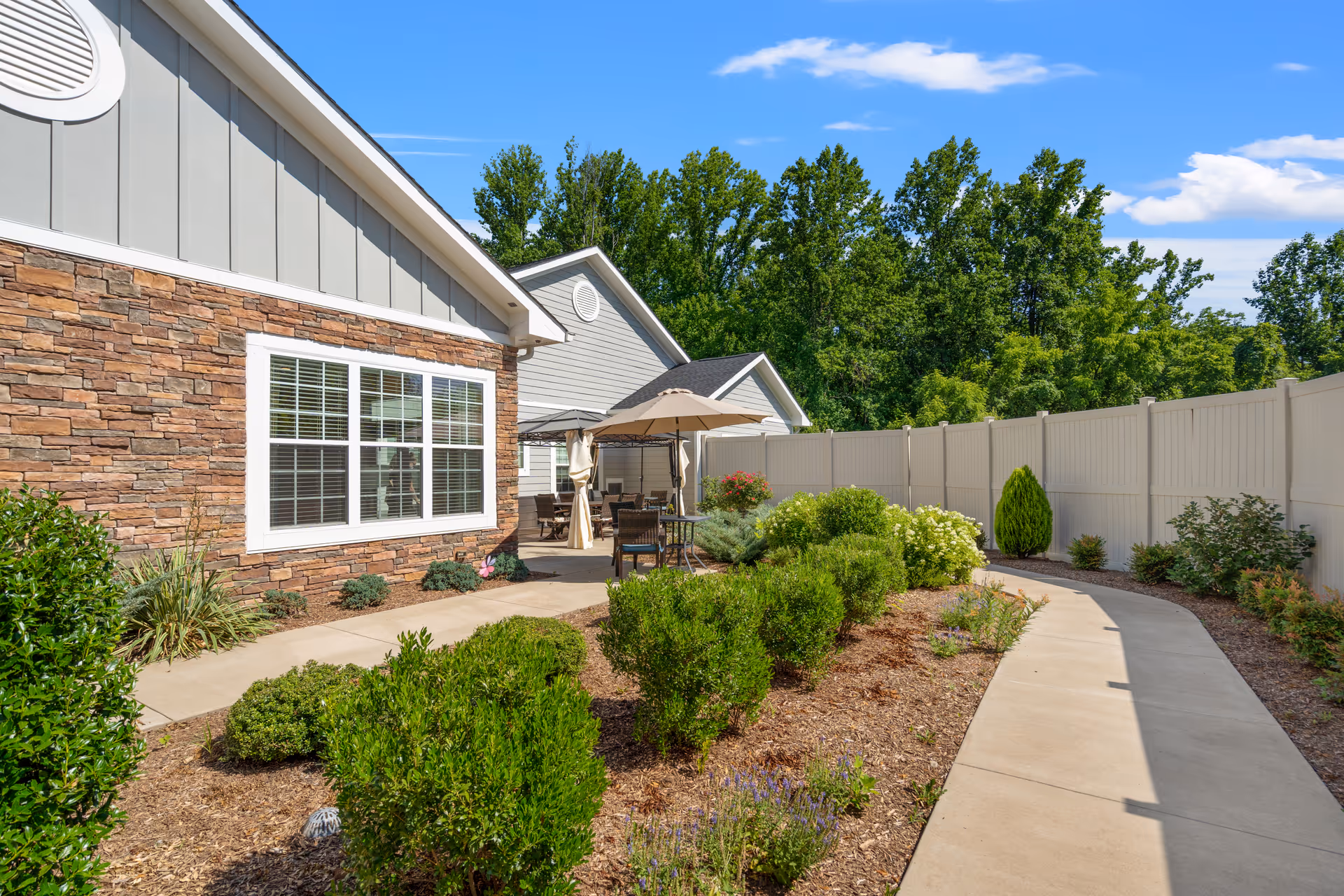 Outdoor patio area at Poet's Walk Warrenton, A Memory Care Community, featuring a paved walkway, green shrubs and plants, a stone and siding building exterior with large windows, patio tables with umbrellas, and a white privacy fence with trees in the background under a blue sky.