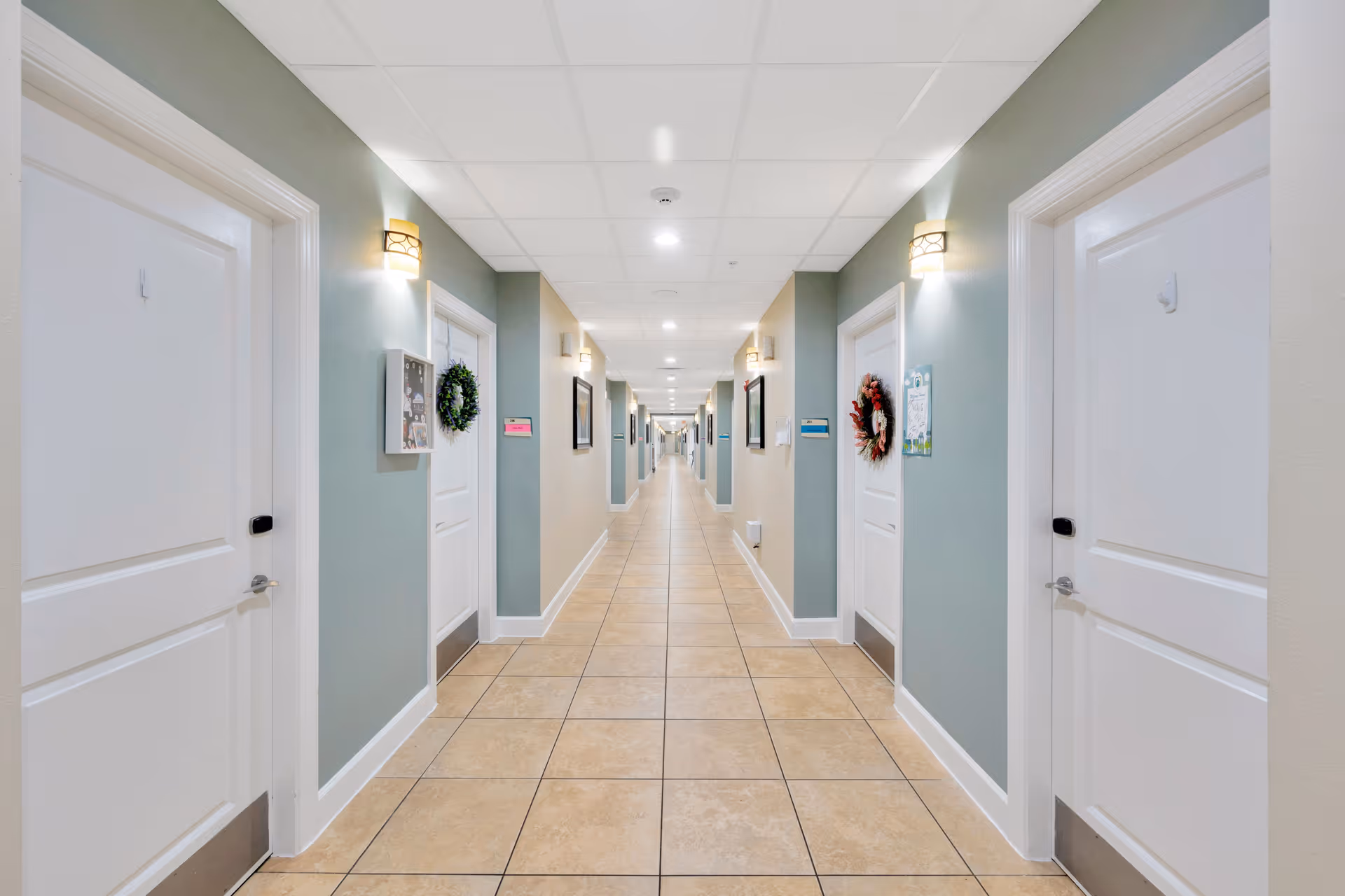 A long, well-lit hallway in a senior living facility with tiled floors and light green walls. White doors line both sides of the corridor, some decorated with wreaths and small signs. Wall-mounted lights illuminate the hallway evenly.
