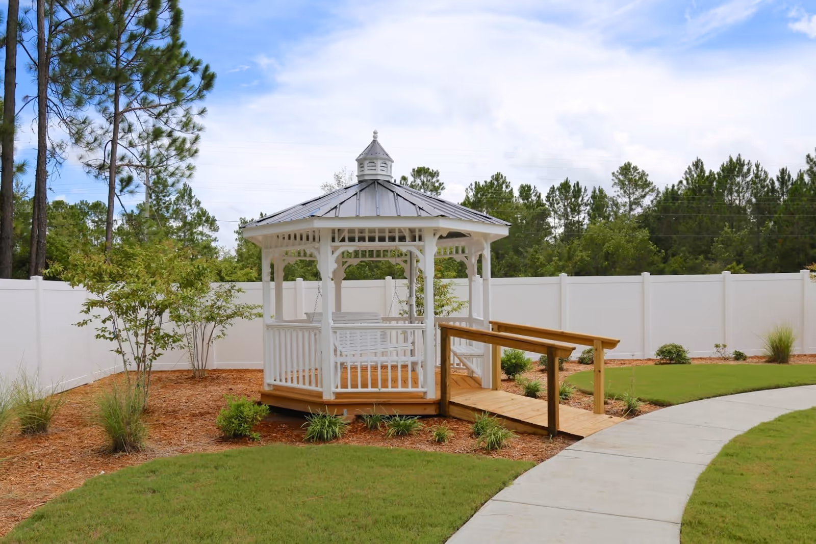 A white gazebo with a metal roof and a wooden ramp is situated in a landscaped garden area with green grass, small plants, and mulch. The gazebo has a white swing inside and is surrounded by a white fence with tall trees in the background under a partly cloudy sky.