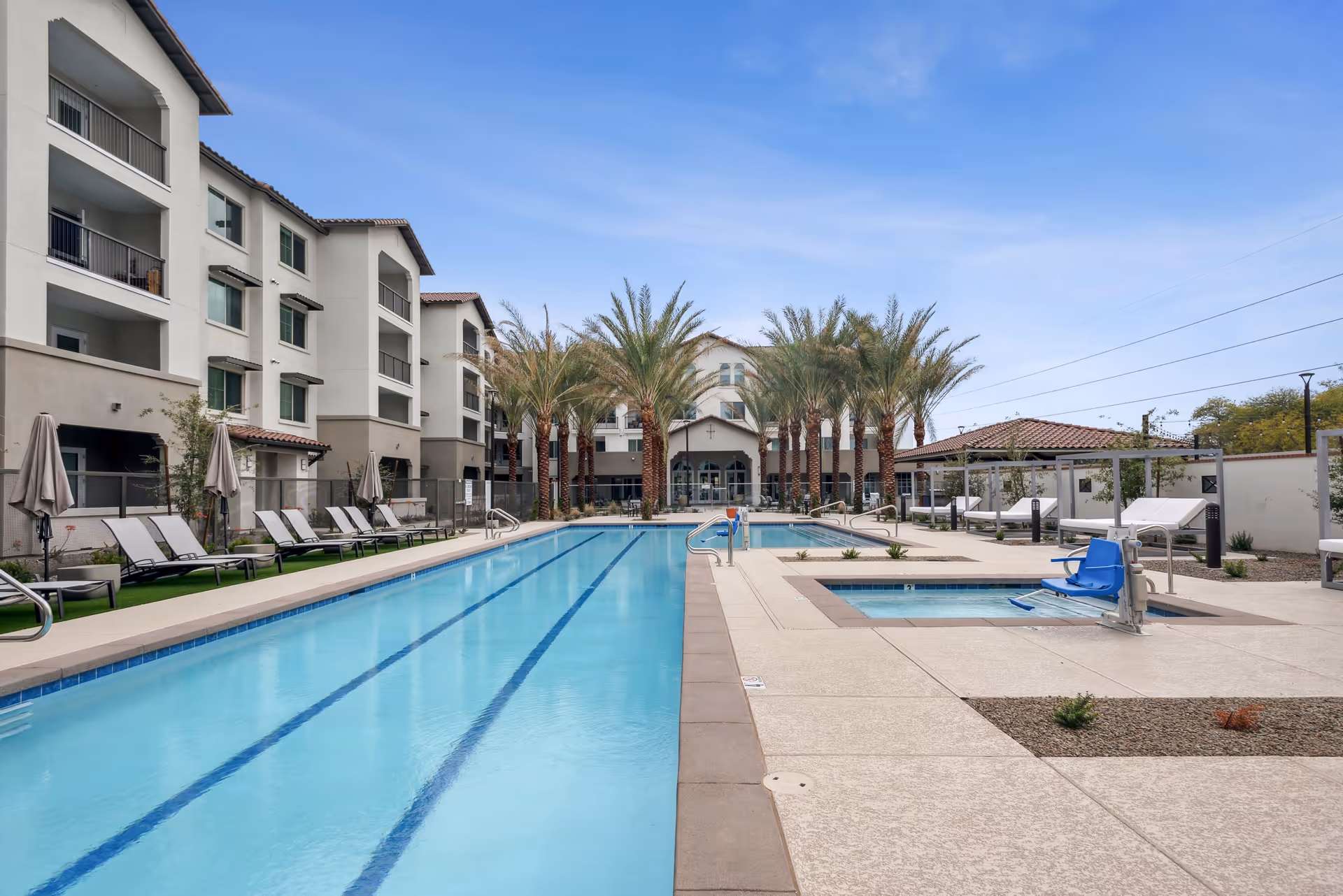 Outdoor swimming pool area at Overture North Scottsdale with lounge chairs and umbrellas along the poolside, palm trees lining the far end, and a multi-story building in the background under a clear blue sky.
