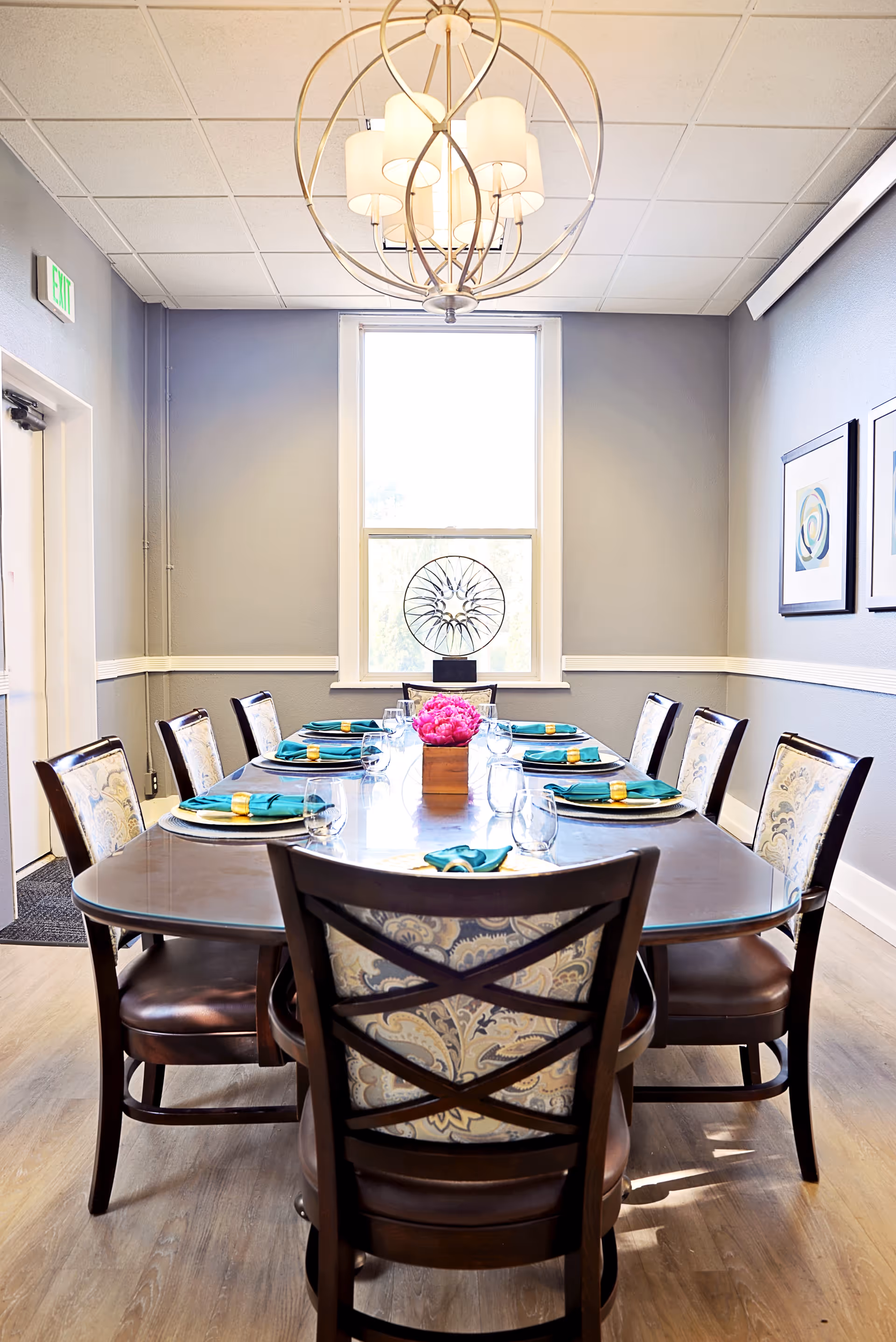 A dining room with a long wooden table set for eight people. Each place setting has a teal napkin with a yellow napkin ring, a plate, and a glass. A decorative centerpiece with pink flowers is in the middle of the table. The room has light gray walls, a large window at the far end, two framed artworks on the right wall, and a modern chandelier hanging from the ceiling.