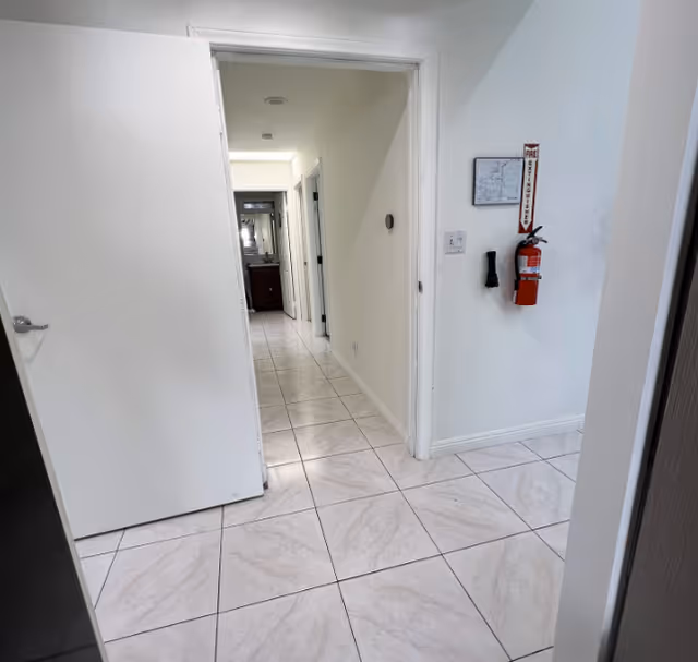 A clean, tiled hallway in an assisted living facility with white walls and an open door on the left. A fire extinguisher is mounted on the right wall with a sign above it. The hallway leads to other rooms with visible doorways and a glimpse of a sink and mirror at the far end.