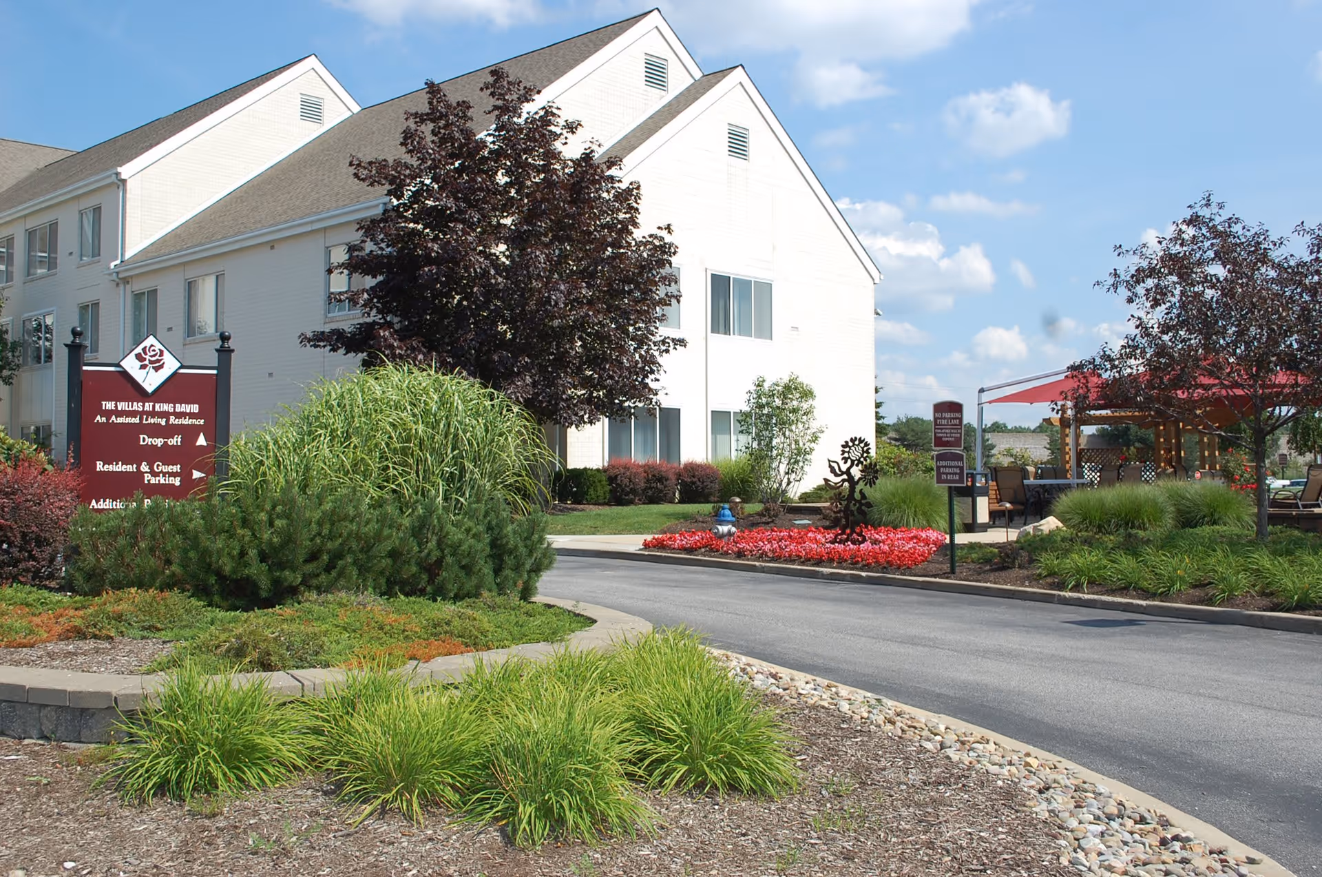 Exterior view of The Villas at King David, an assisted living residence, showing a white two-story building with a landscaped driveway, green bushes, trees, and a red sign indicating drop-off, parking, and additional parking directions. There is also a seating area with red umbrellas visible on the right side.