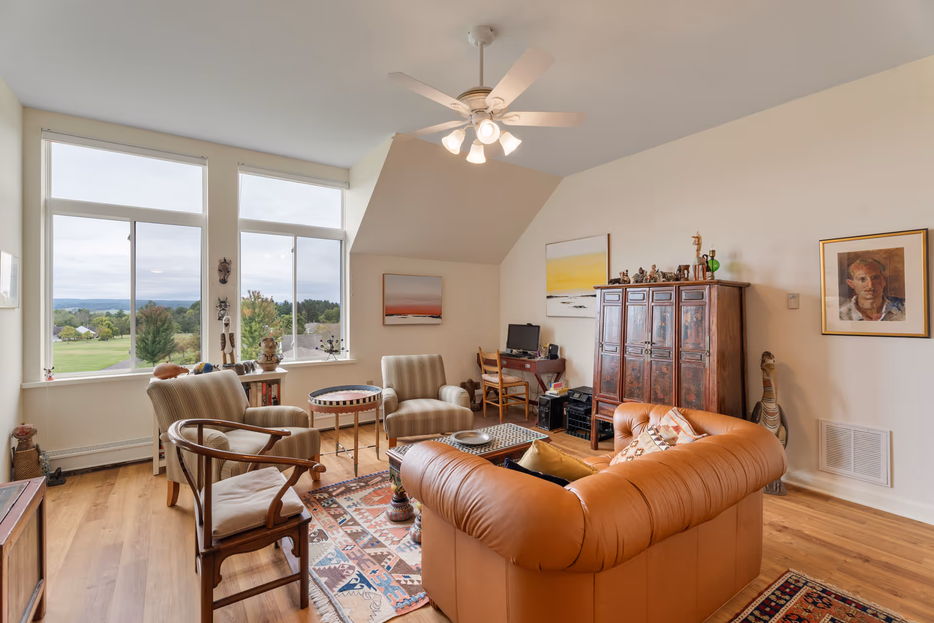 A cozy living room with a large brown leather sofa, two striped armchairs, a wooden chair, and a small round table. The room has large windows showing a green outdoor landscape, wooden flooring, a ceiling fan with lights, and various artworks and decorative items on the walls and furniture.