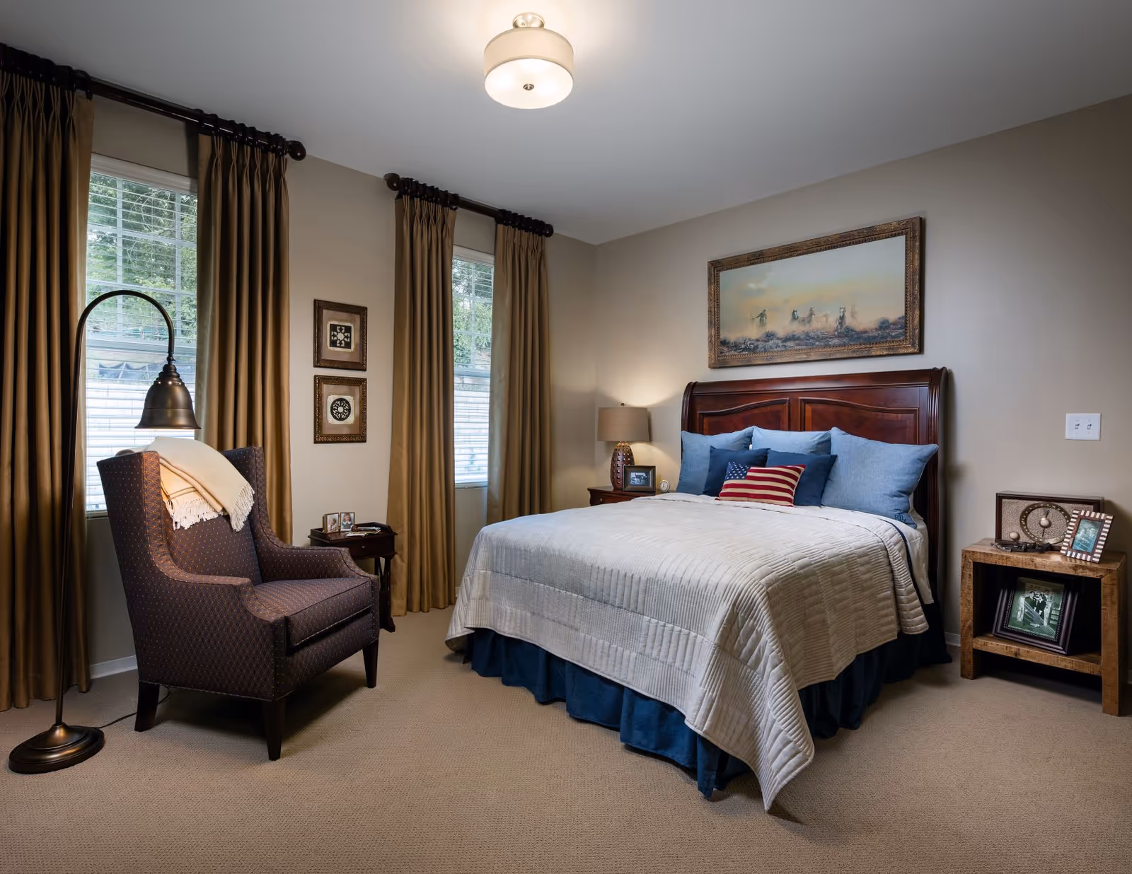 Cozy, well-lit bedroom with a made bed, decorative pillows and wooden headboard, an armchair, bedside tables, lamps, and long curtains.