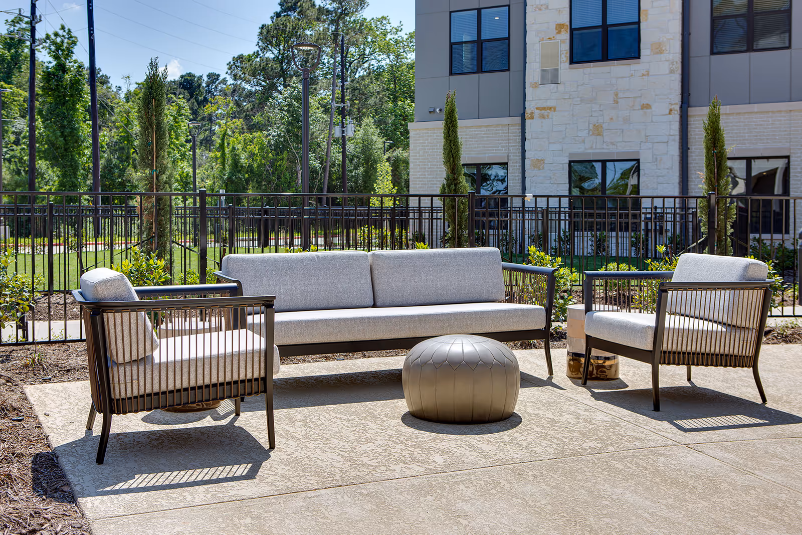 Outdoor patio area with modern cushioned seating including a sofa and two armchairs arranged around a round metallic ottoman. The area is paved and surrounded by a black metal fence with greenery and trees in the background. A building with stone and brick exterior is visible behind the fence.