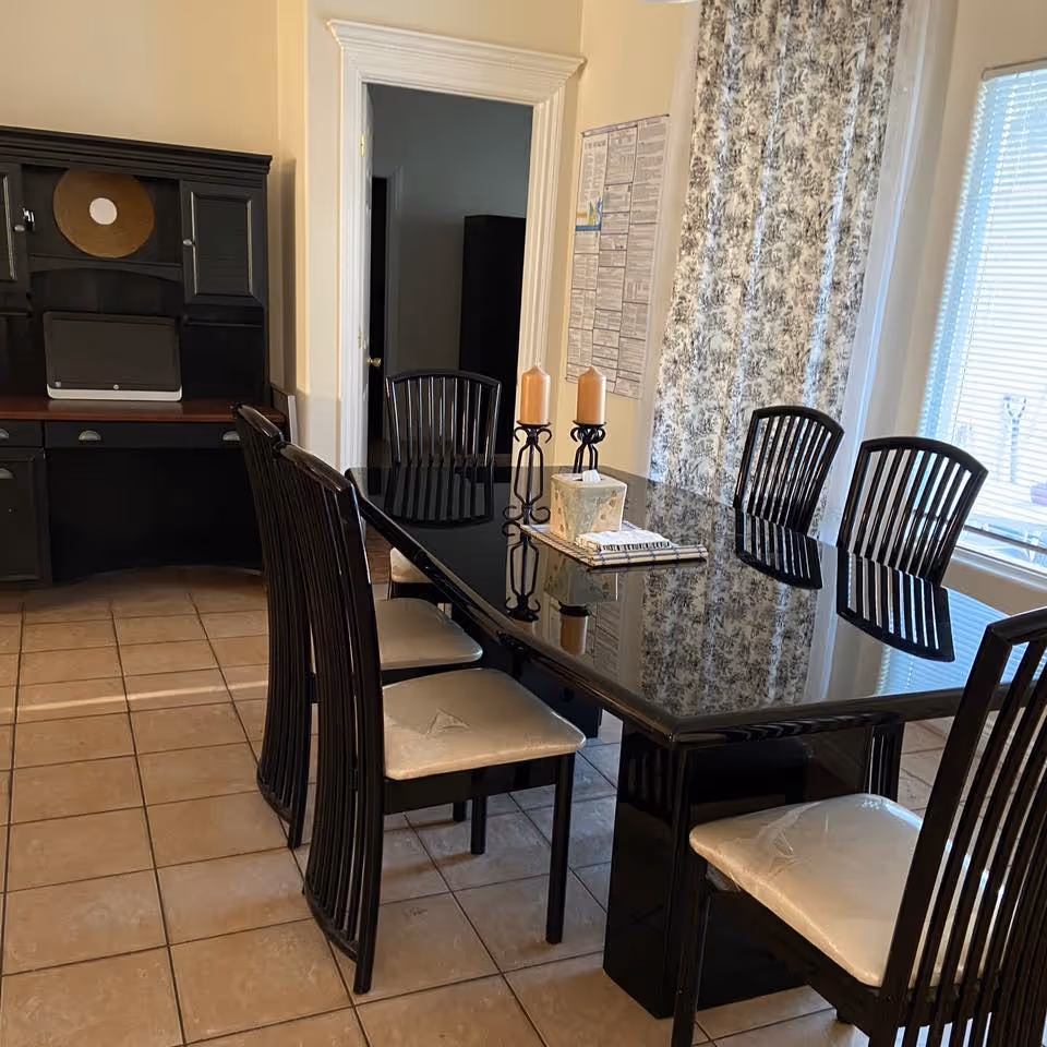 A dining room with a black glossy rectangular table surrounded by six black wooden chairs with light-colored cushions. On the table are two candles on black holders, a tissue box, and a book. The room has tiled flooring, a window with patterned curtains, and a dark wooden desk with a laptop in the background.