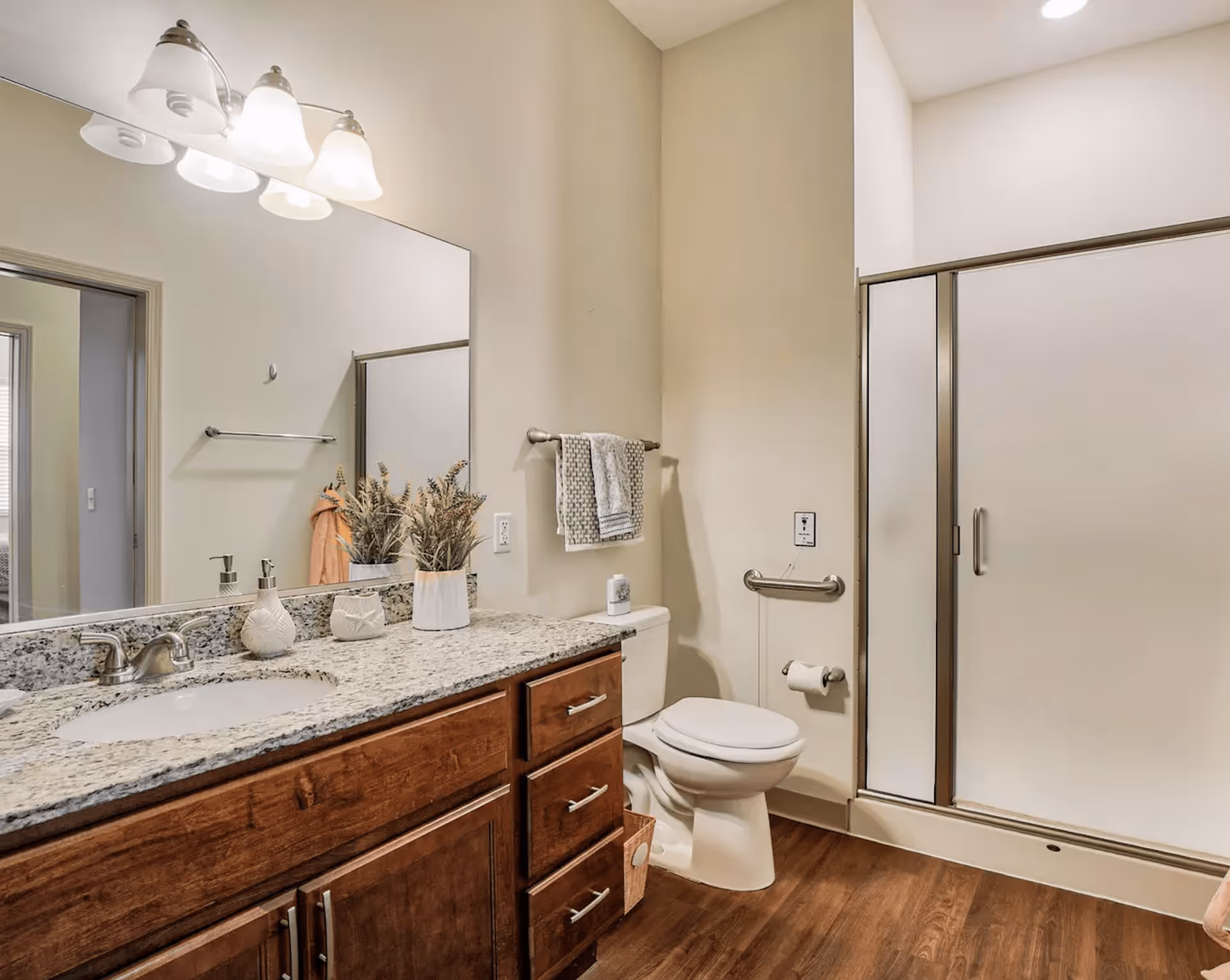 A clean and well-lit bathroom featuring a granite countertop with a sink, a large mirror with three light fixtures above it, a toilet with a towel rack and handrail beside it, and a glass-enclosed shower. The floor is wooden, and there are decorative items on the countertop including a plant and soap dispensers.