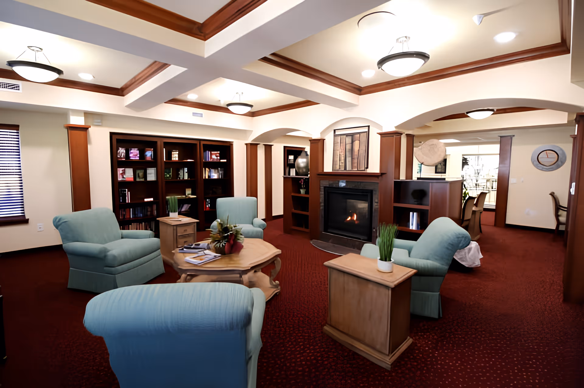A cozy common room with light blue armchairs arranged around a wooden coffee table, a fireplace, bookshelves, and recessed ceiling lighting.