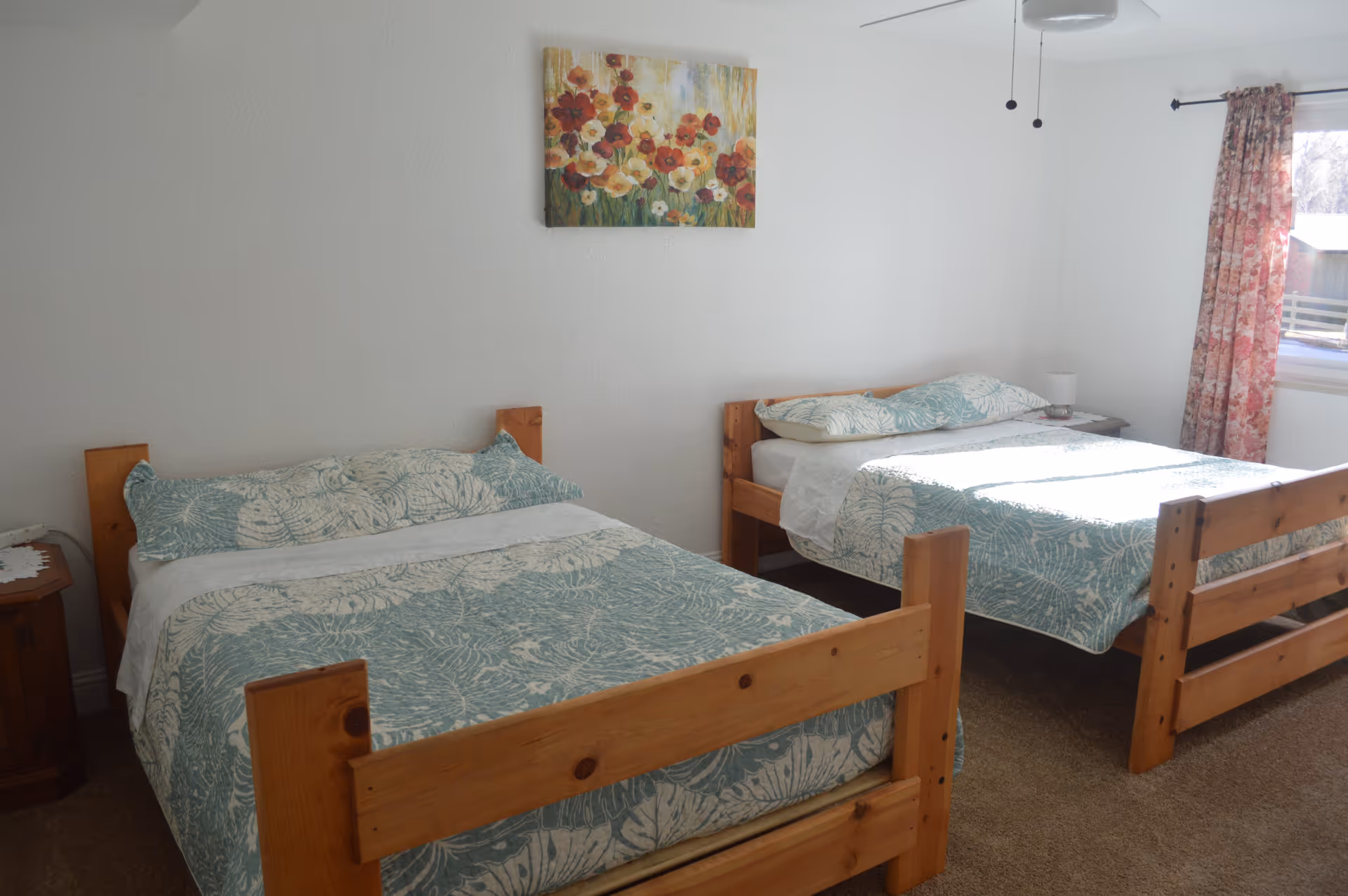 A bedroom with two wooden beds, each made with matching blue and white patterned bedding. There is a small wooden nightstand with a lamp between the beds. A window with floral curtains allows natural light to brighten the room. A colorful floral painting hangs on the white wall above the beds.
