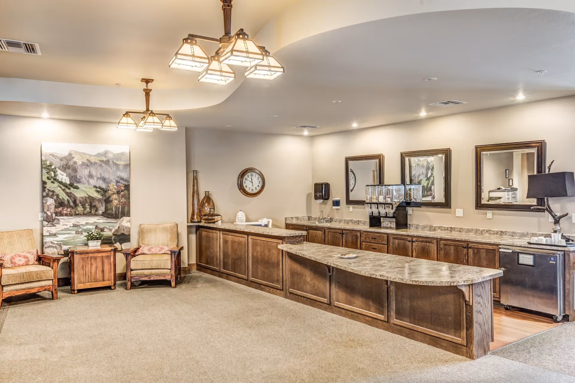 A spacious interior room with a long marble countertop and wooden cabinets underneath. On the countertop, there are beverage dispensers and a small sink. Two mirrors and a framed painting hang on the wall above the counter. To the left, there are two cushioned armchairs with wooden frames, a small wooden side table between them, and a large landscape painting behind them. The room is softly lit with ceiling lights and has a carpeted floor.