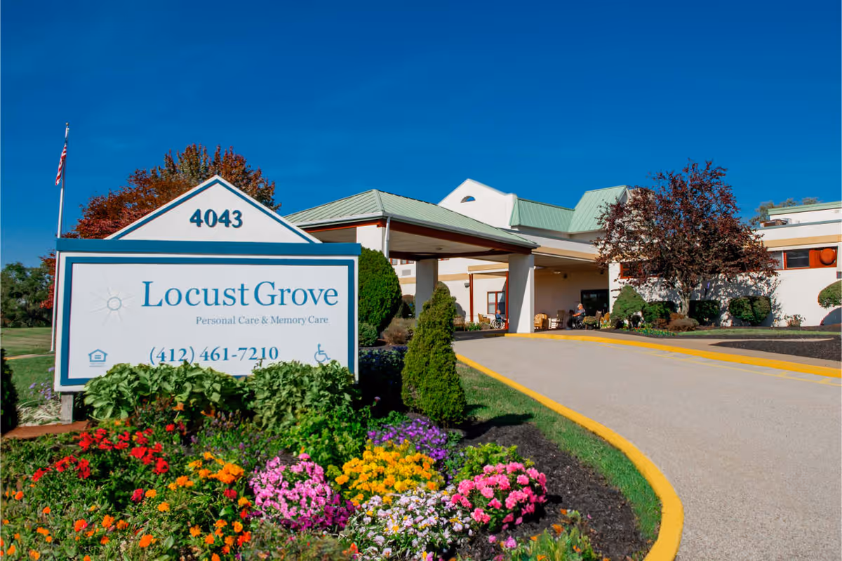 Exterior view of Locust Grove Personal Care & Memory Care facility with a large sign displaying the name, phone number, and address 4043. The building has a covered entrance, green roof, and is surrounded by landscaped gardens with colorful flowers and trimmed bushes under a clear blue sky.