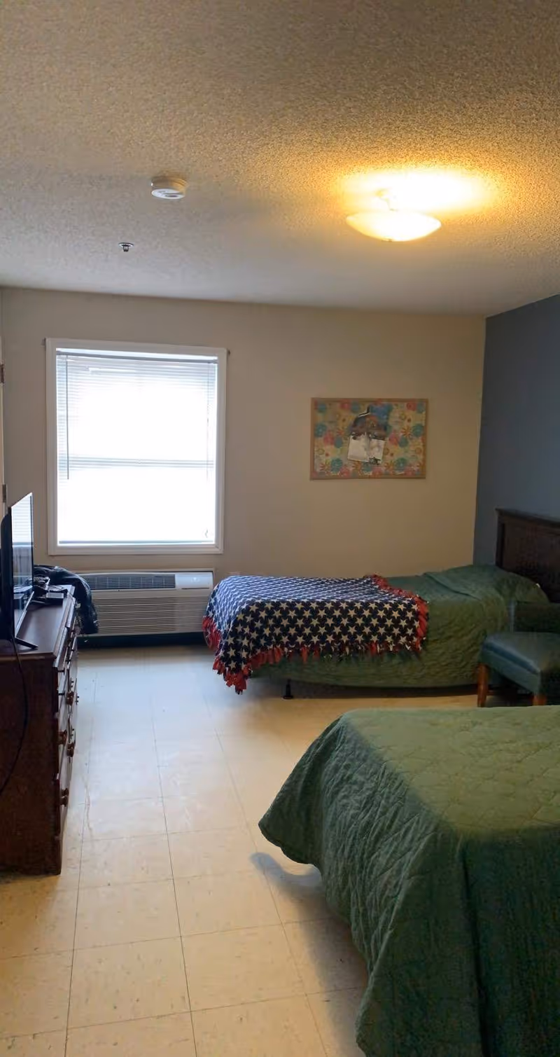 A simple bedroom with two single beds covered in green bedspreads. One bed has a star-patterned blanket draped over it. There is a wooden dresser with a TV on top, a window with blinds, a wall-mounted air conditioning unit below the window, and a colorful bulletin board on the wall.