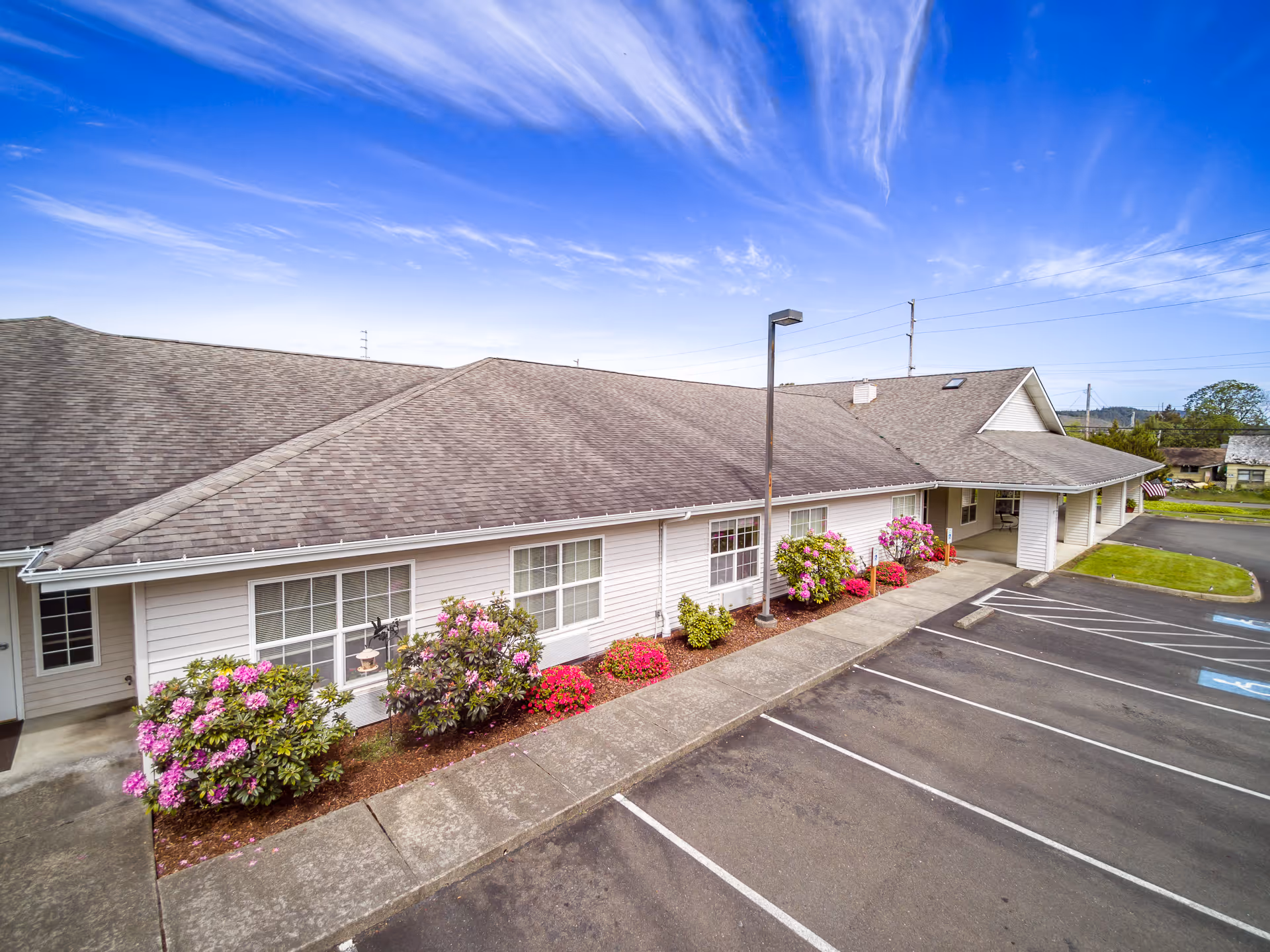 Exterior view of a single-story building with a gray shingled roof and white siding. The building is surrounded by a sidewalk and landscaped with flowering bushes. There is a parking lot with marked spaces, including handicapped spots, adjacent to the building. The sky is clear with some wispy clouds.