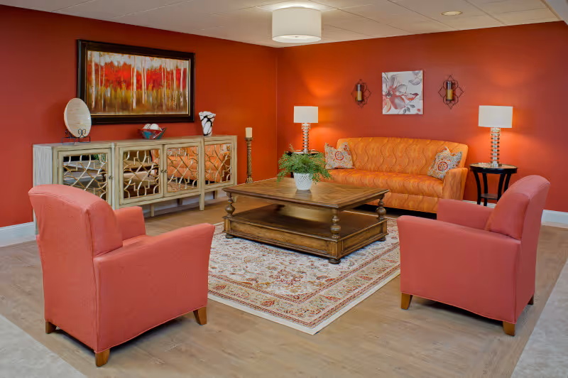 A cozy living room with red walls featuring an orange patterned sofa, two red armchairs, a wooden coffee table on a decorative rug, a sideboard with mirrored doors, and wall art including a painting of trees and a floral print. Two table lamps and wall sconces provide warm lighting.