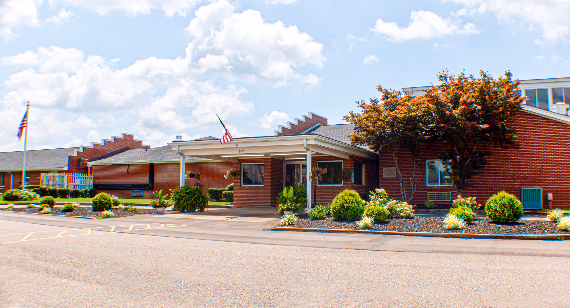 Front entrance of a single-story brick healthcare and rehabilitation center with a covered drop-off, landscaped beds, and American flags.