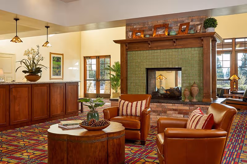 A cozy senior living lounge with leather armchairs around a wooden coffee table facing a tiled fireplace and a reception desk.