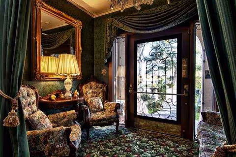 Luxurious, vintage-style living room with patterned carpet, upholstered chairs, ornate mirror, lamp, and wrought-iron doorway.