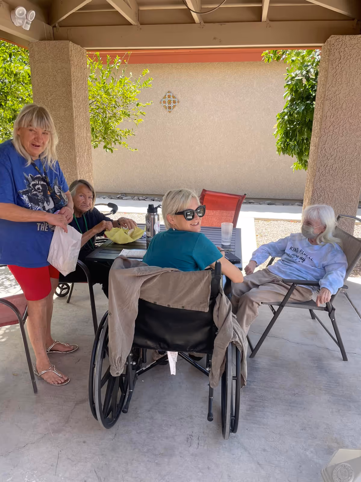 Four elderly women sitting and standing around a table under a covered outdoor patio. One woman is in a wheelchair wearing sunglasses and smiling at the camera. Another woman is standing wearing a blue shirt and red shorts. The other two women are seated, one wearing a face mask. There are plants and a beige wall in the background.