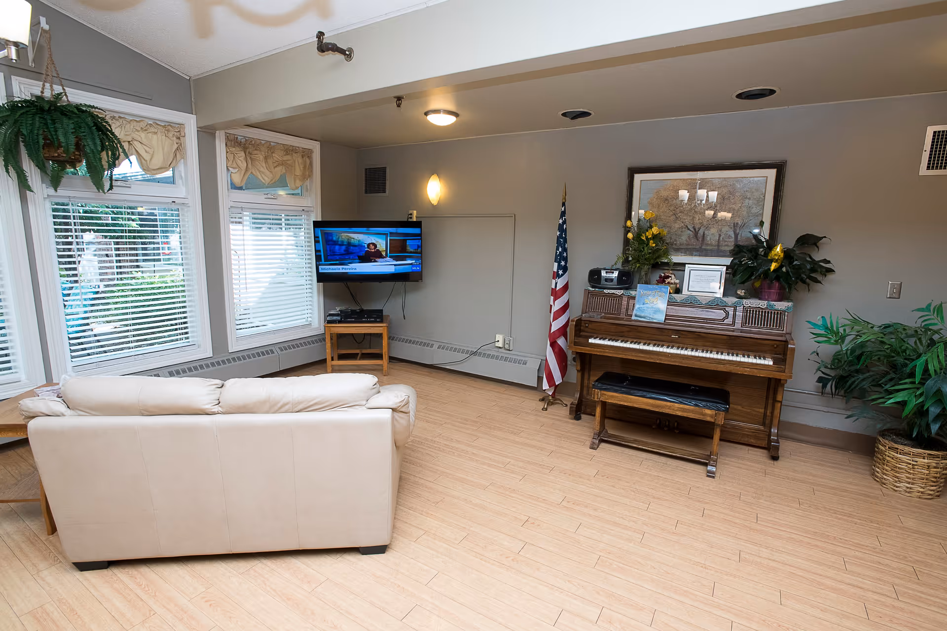 A common room with a beige sofa facing a wall-mounted TV and an upright piano decorated with plants and an American flag.